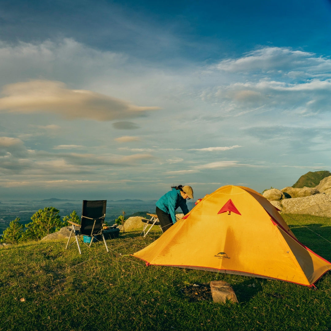 Person setting up a yellow tent on a grassy hill with a scenic view and blue sky.